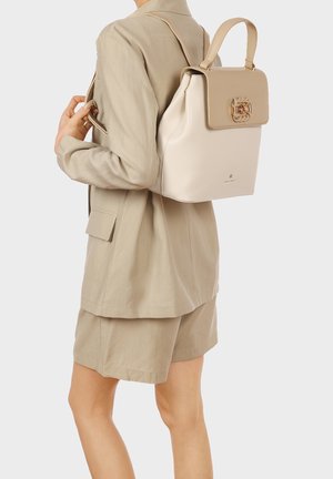 Persona che indossa un blazer beige e pantaloncini, con uno zaino in pelle color crema e marrone chiaro, con fibbia dorata, portato sulla spalla.