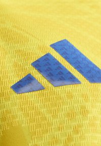 Trois bandes bleues du logo Adidas sur un fond en tissu jaune texturé.