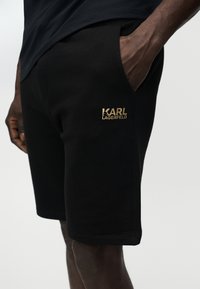 Shorts noirs avec une main dans la poche et le logo doré "Karl Lagerfeld" sur la cuisse, portés avec une chemise noire.