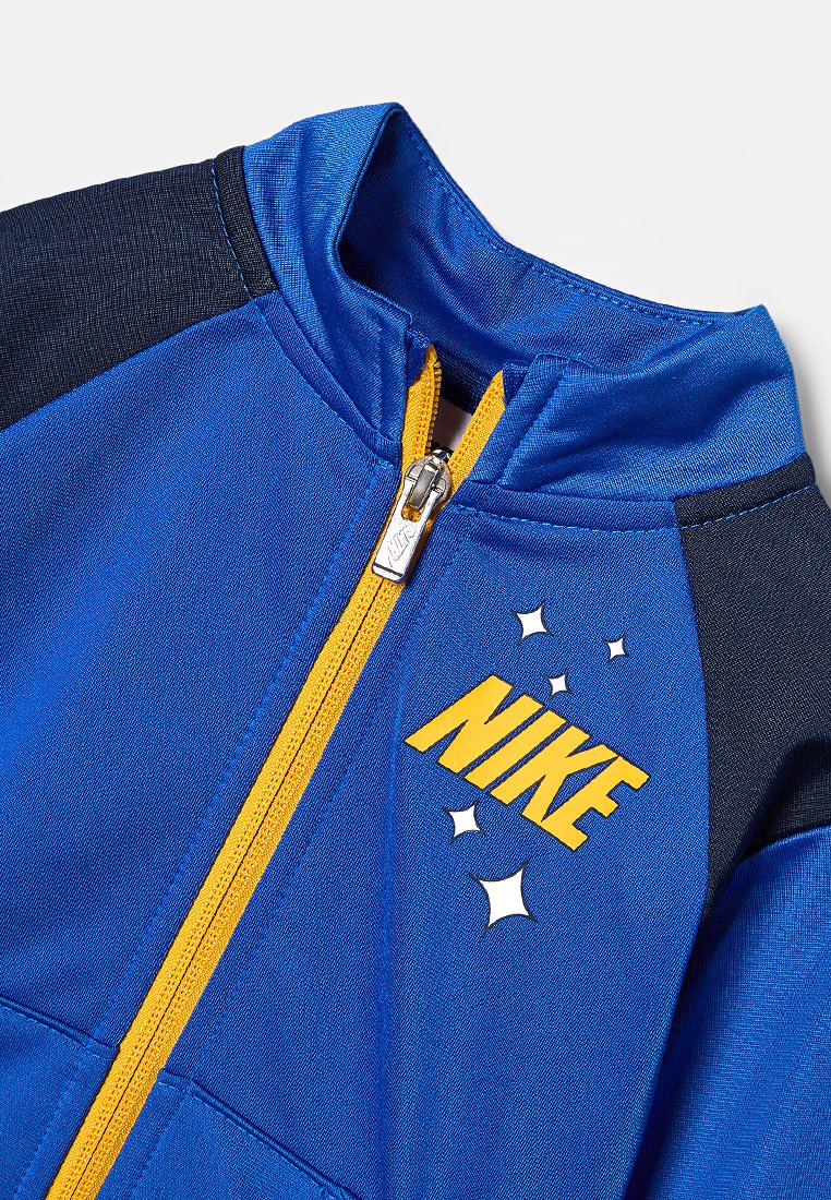 Giacca sportiva blu con colletto alto, cerniera gialla e logo "NIKE" in giallo. Presenta dettagli neri e una stella sul petto.