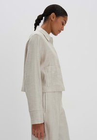 Beige, verkort linnen jasje met een kraag en een zijzak. Heeft een losse pasvorm en lange mouwen, getoond vanuit de zijkant.