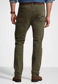 Pantalones de algodón verde oliva con un diseño de pierna recta. Incluyen bolsillos traseros, una etiqueta de cuero marrón y detalles de costura visibles.