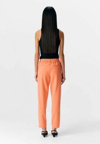 Haut sans manches noir associé à un pantalon tailleur corail orange avec une taille élastique et deux poches arrière, porté avec des talons noirs.