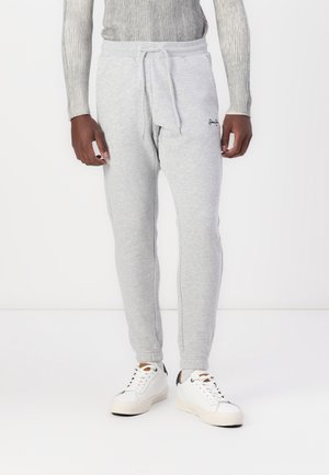 Homme portant un pantalon de jogging gris clair avec cordon de serrage et des baskets blanches sur un fond blanc uni.