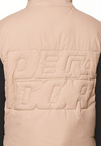 Padded jacket v svetlej béžovej farbe s čiernymi rukávmi, na zadnej strane s embosovaným logom. Švy sú viditeľné, čo dodáva textúrovaný vzhľad.