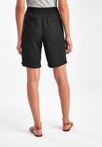 Vrouw draagt zwart, mid-lengte korte broek met elastische tailleband en achterzakken, gecombineerd met bruine sandalen met gesp, staand op een witte achtergrond.