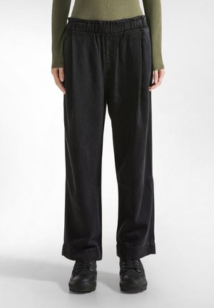 Pantaloni neri a gamba larga con elastico in vita, plissettature e risvolti. Realizzati in un tessuto strutturato, abbinati a sneakers nere.