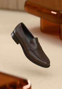 Brune lær loafers med en syet detalj og en glatt overflate, med en svart såle. Plassert mot en beige bakgrunn med amberakenter.