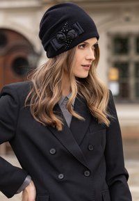 Gorro de lana negro con acabado texturizado, que presenta acentos florales negros y una cinta. Combinado con un blazer negro de doble botonadura y una camisa gris.