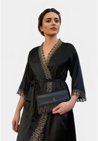 Bata de satén negra con un borde de encaje de diseño a lo largo del escote y las mangas, atada a la cintura, combinada con un bolso de mano negro que presenta un logo de corona.