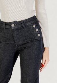 Lähikuva henkilöstä, jolla on yllään mustat farkut, joissa on koristeelliset hopeanväriset napit taskussa, ja vaaleanbeige ribattu pitkähihaista paitaa.