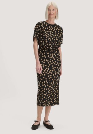Vrouw met stijl wit haar draagt een zwarte midi-jurk met beige polkadots en zwarte platte schoenen met enkelbandjes, staand tegen een effen achtergrond.