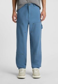 Blauwe cargobroek met een ruime pasvorm, voorzien van grote zakken en metalen drukknopen, gecombineerd met beige sneakers. Glad stof, rechte pijpen.