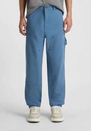 Blue cargo pants with a loose fit, featuring large pockets and metal snaps, paired with beige sneakers. Smooth fabric, straight leg design.