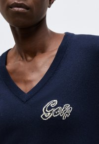 Κοντινό πλάνο ατόμου που φορά μπλε σκούνο πουλόβερ με V λαιμόκοψη και λευκό κεντητό κείμενο "Golf" στο αριστερό στήθος, με απλό ανοιχτό φόντο.