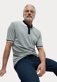 Ljust grå pikétröja med texturerad tyg, marinblå krage och manschettdetaljer, bärs med blå jeans, modell sitter med händerna på låret.