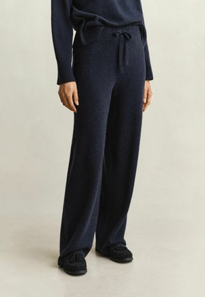 Personne portant un pantalon large en maille bleu marine foncé avec une taille à cordon et un pull assorti foncé, associé à des chaussures noires sans lacets.