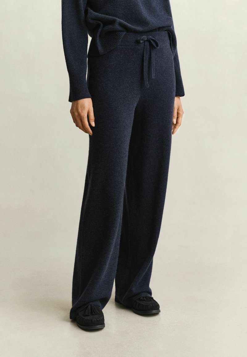 Personne portant un pantalon large en maille bleu marine foncé avec une taille à cordon et un pull assorti foncé, associé à des chaussures noires sans lacets.