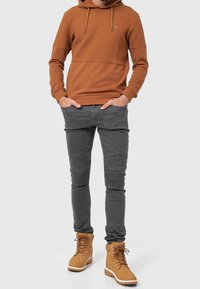 Mężczyzna w brązowej, teksturowanej hoodies, dopasowanych szarych spodniach z detalami na kolanach i beżowych butach na sznurówki stoi z rękami w kieszeniach.
