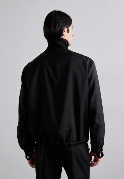 Black jacket with a high collar, elastic waistband, and cuffs. Smooth texture, relaxed fit, and rounded shoulder design.