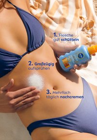 Sunscreen bottle with an orange cap and blue body, instruction text displayed, applied on skin against a light-colored towel backdrop.