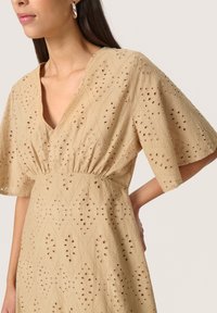 Beige bomullsklänning med diamantmönster och hålstickade detaljer, med en v-ringning och vida ärmar, accentuerad av en rynkad midja.