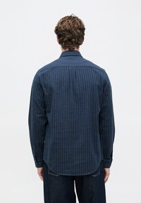 Hombre con cabello castaño rizado, vistiendo una camiseta de manga larga a rayas azul oscuro y pantalones oscuros, de pie de espaldas contra un fondo blanco.