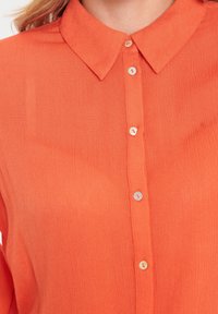 Eine Frau trägt ein leuchtend oranges Hemd mit Knopfleiste und spitzem Kragen, das unter dem Kinn endet.