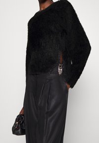 Kvinna klädd i en svart fluffig tröja med sidoklyfta och dekorativ brosch, i kombination med högmidjade svarta plisserade byxor, hållande en svart väska.