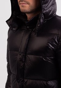 Černá prošívaná bunda s lesklým povrchem. Má vysoký límec a kapuci, které se zapínají na knoflíky a přezky.