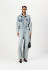 Lys blå denimjakke med to brystlommer og matchende vide bukser. Sort håndveske og spisse sko fullfører antrekket.