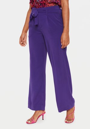 Femme portant un pantalon large taille haute violet avec une ceinture nouée et des sandales à talons roses, debout devant un fond clair uni.
