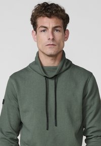 Joven con cabello rizado que lleva una sudadera verde con textura y cordones negros, mirando hacia adelante sobre un fondo liso.
