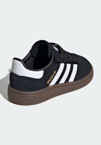 Black suede sneaker with white leather accents, three horizontal stripes, gold branding, and a textured brown rubber sole.