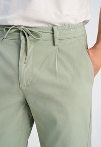 Pantaloni leggeri di colore verde chiaro, realizzati in tessuto morbido, con fascia elastica in vita, chiusura con lacci, tasche laterali e una leggera texture.