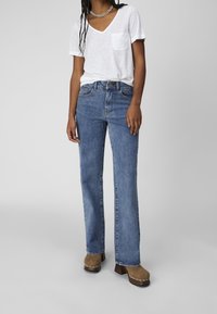 Person iført en hvid V-hals-t-shirt med lomme, blå lige ben jeans og brune klipklapper med metalnitter på en grå baggrund.