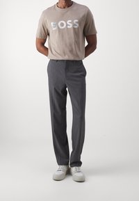Muž stojící s rukama za zády, oblečený v béžovém tričku "BOSS", šedých kalhotách a bílých teniskách na jednobarevném světlém pozadí.