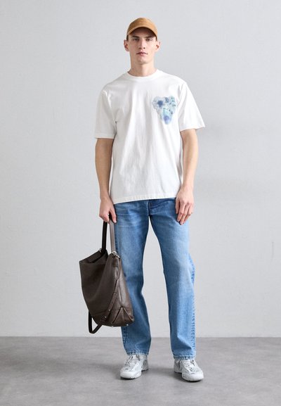 Camiseta de algodón blanca con un diseño floral colorido en el pecho, combinada con unos pantalones de denim azul claro. El modelo sostiene un bolso de hombro de cuero marrón.
