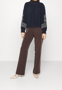 Marinebluse mit floralen Stickereien an den Ärmeln, kombiniert mit braunen weit geschnittenen Hosen und nude-farbenen High Heels. Glatte Stoffe überall.