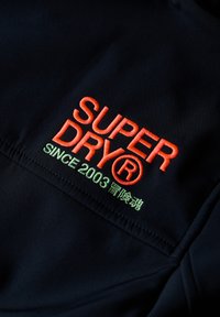 Broderi på mørkt stof med teksten "SUPER DRY®" i orange og "SINCE 2003" med japanske tegn i grønt.
