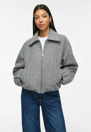 Donna che indossa una giacca di lana grigia con zip e colletto e jeans blu, in piedi con le mani nelle tasche davanti a uno sfondo bianco uniforme.