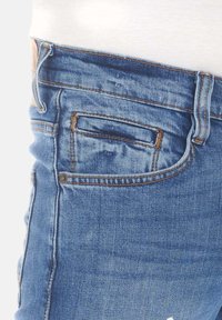 Los jeans de denim azul presentan un diseño ajustado con una cintura de altura media, costuras intrincadas y un pequeño bolsillo delantero. La tela tiene una textura ligeramente desgastada.