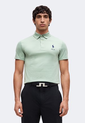 Homme portant un polo vert clair avec un logo bleu marine et un pantalon noir avec une ceinture noire avec boucle argentée, debout, les bras détendus.