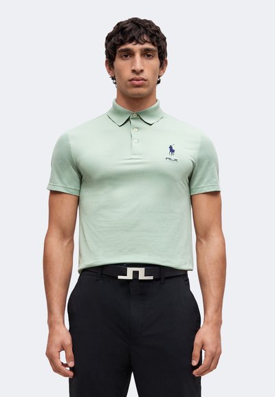 Homme portant un polo vert clair avec un logo bleu marine et un pantalon noir avec une ceinture noire avec boucle argentée, debout, les bras détendus.