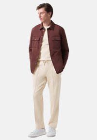 Braune gepolsterte Jacke mit Brusttaschen, beiger Pullover, hellbeige Hose mit seitlichen Paspeln und weiße Sneaker. Allgemein glatte Textur.