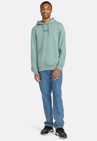 Mint green hoodie with kangaroo pocket and logo, paired with blue denim jeans and black sneakers with white accents. Casual fit.