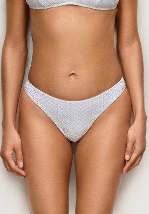 Kobieta ubrana w biały teksturowany dół bikini i pasujący top, stojąca na tle jednolitego jasnego tła, ręce swobodnie opadające wzdłuż ciała.