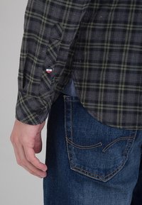 Langsleeve geruite shirt in donkergroen en marineblauw met knoopmanchetten, voorzien van een klein, multicolor logo patch; gedragen met blauwe jeans.
