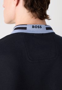 Črna polo majica z svetlo modrim ovratnikom, ki ima črne stripe. Na ovratniku je izpisano logo "BOSS". Ima gladko teksturo in prilegajoč kroj.