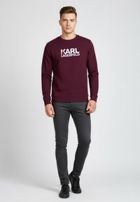 Bordeauxfarbenes Sweatshirt mit dem weißen "KARL LAGERFELD"-Logo, kombiniert mit schwarzen engen Hosen und schwarzen Turnschuhen. Einfaches Rundhalsdesign.